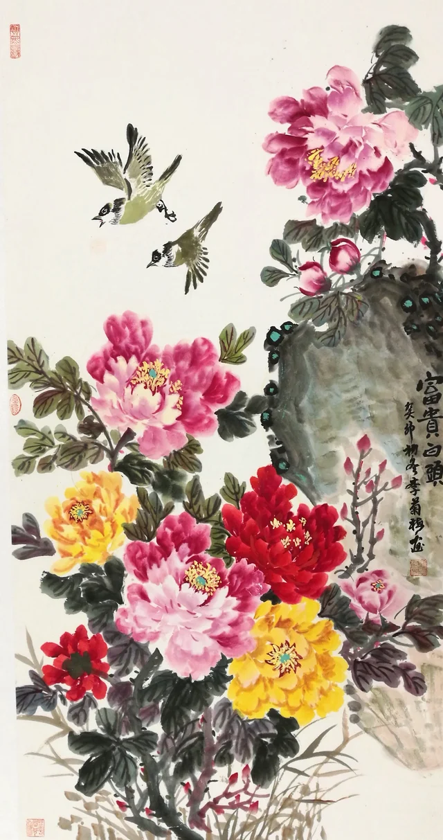 A tall painting of peonies in peach, crimson, yellow and orange rising beside a large mossy rock, with two black-and-white-headed birds swooping down through the empty upper part; calligraphy at the right.