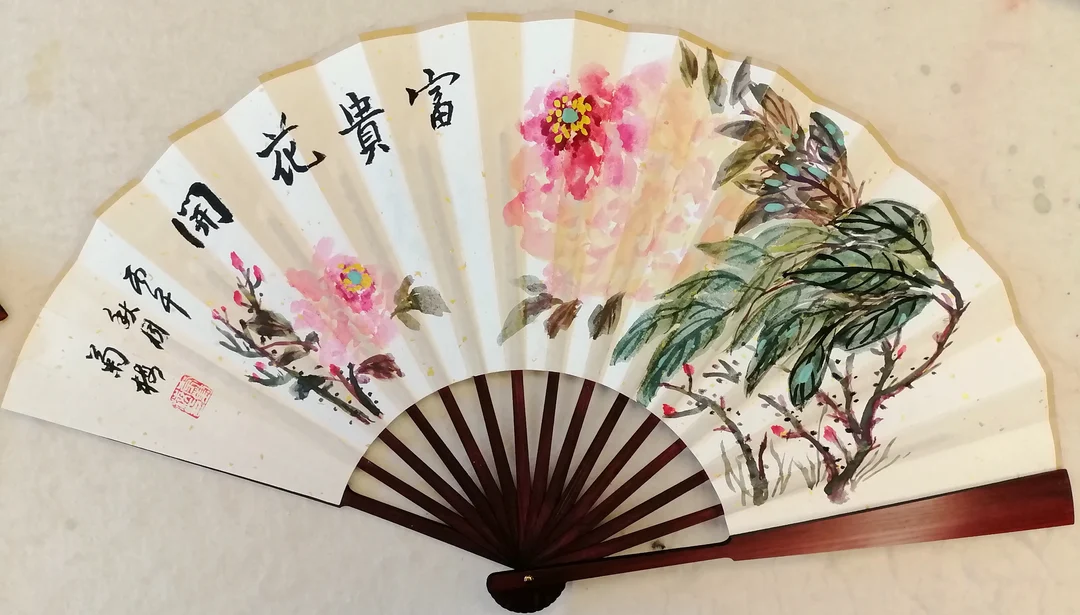 A folding fan painted with two soft pink peonies and a leafy flowering shrub, with four large characters written across the left.